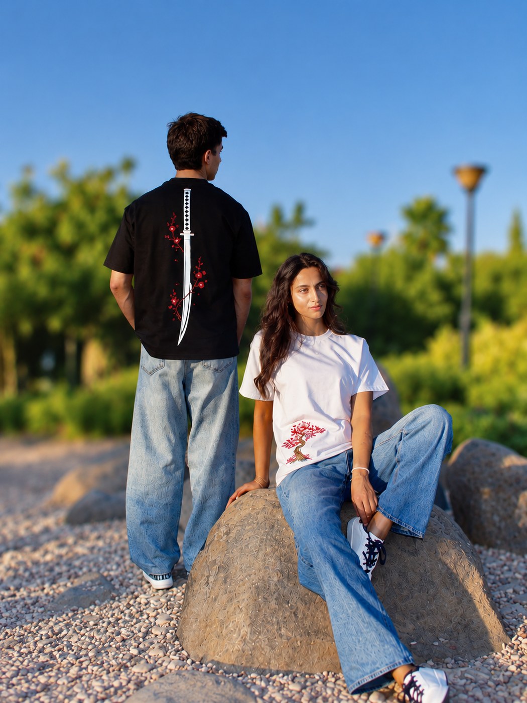 Two models outdoors wearing the IstitchJo Hanabi black tee with embroidered katana design and the Himeno white tee with embroidered blossom tree design
